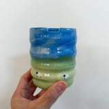 A hand holding a small handmade ceramic cup with a wavy ribbed design, featuring a blue glazed top half and a green glazed bottom half with small white sheep details.
